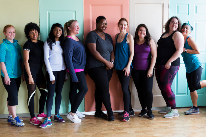 A group of diverse size and ethnicity women in workout clothes lined up against a gym wall